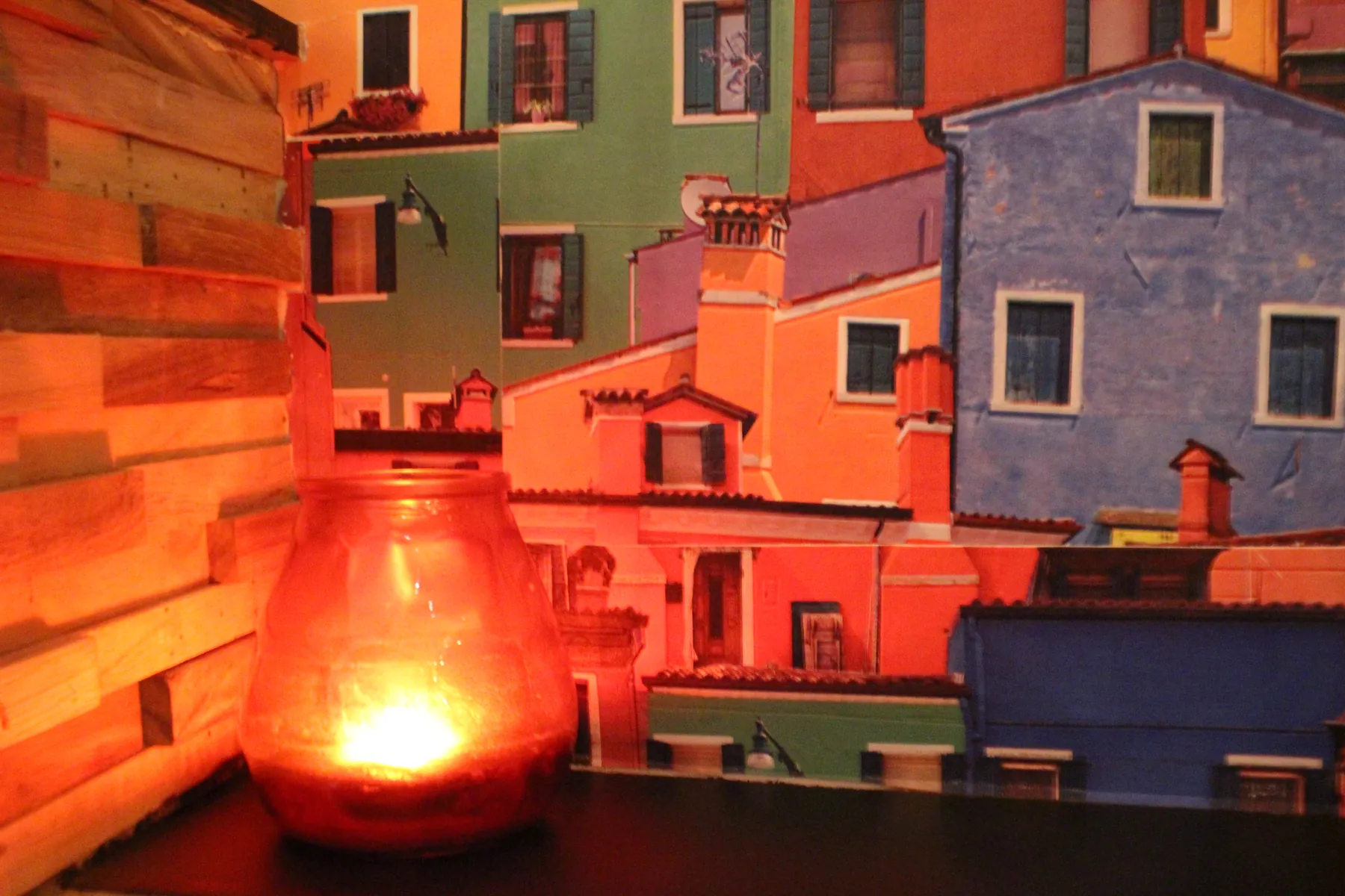 Een brandende kaars in rood glas op tafel, tegen het wandbehang met Italiaanse vissershuisjes